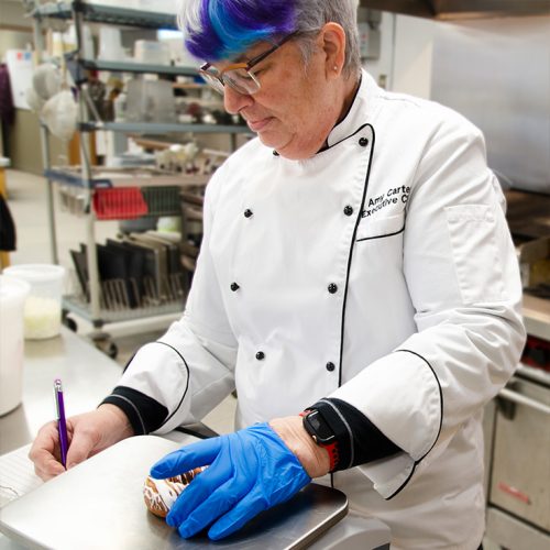 A chef in uniform placing a food item on a scale and making notes in a notebook.